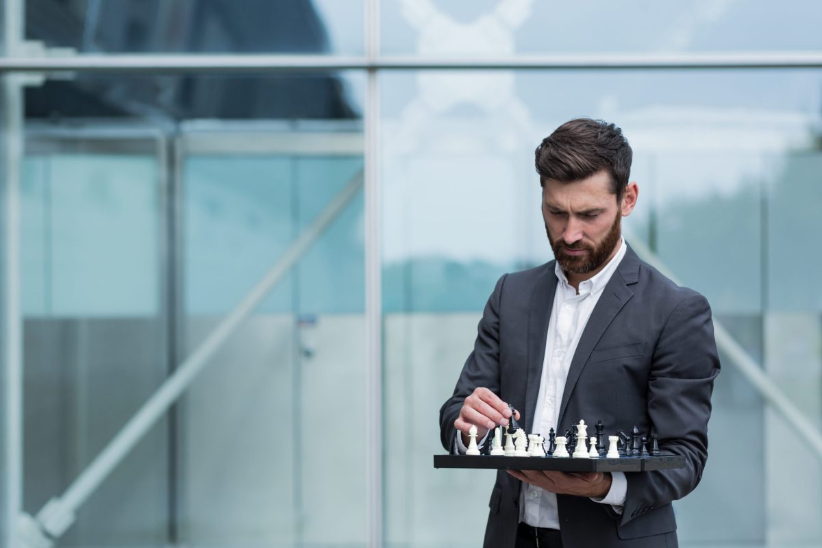 Business broker man standing near office holding board with playing chess, thinking about business strategy Business broker man standing near office holding board with playing chess, thinking about business strategy