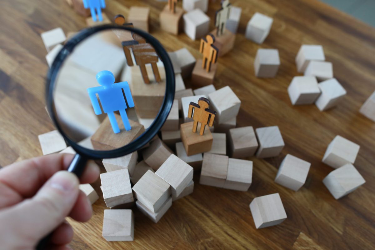Bunch of cubes and miniature people Close-up view of person hand holding magnifying glass and looking through on plastic human model standing on wooden block. Stand out from the crowd concept
