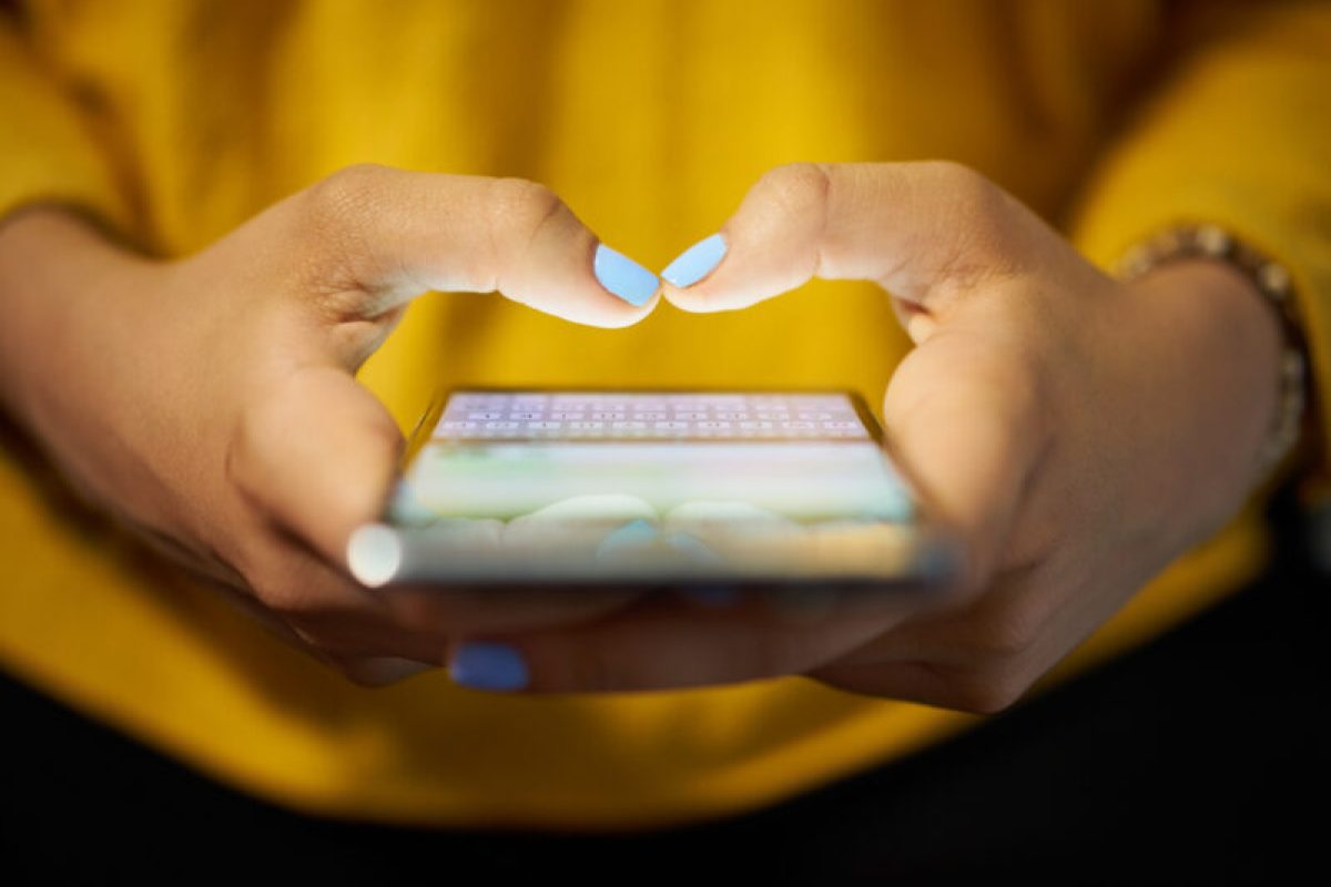 Woman Typing Phone Message On Social Network At Night Young woman using cell phone to send text message on social network at night. Closeup of hands with computer laptop in background