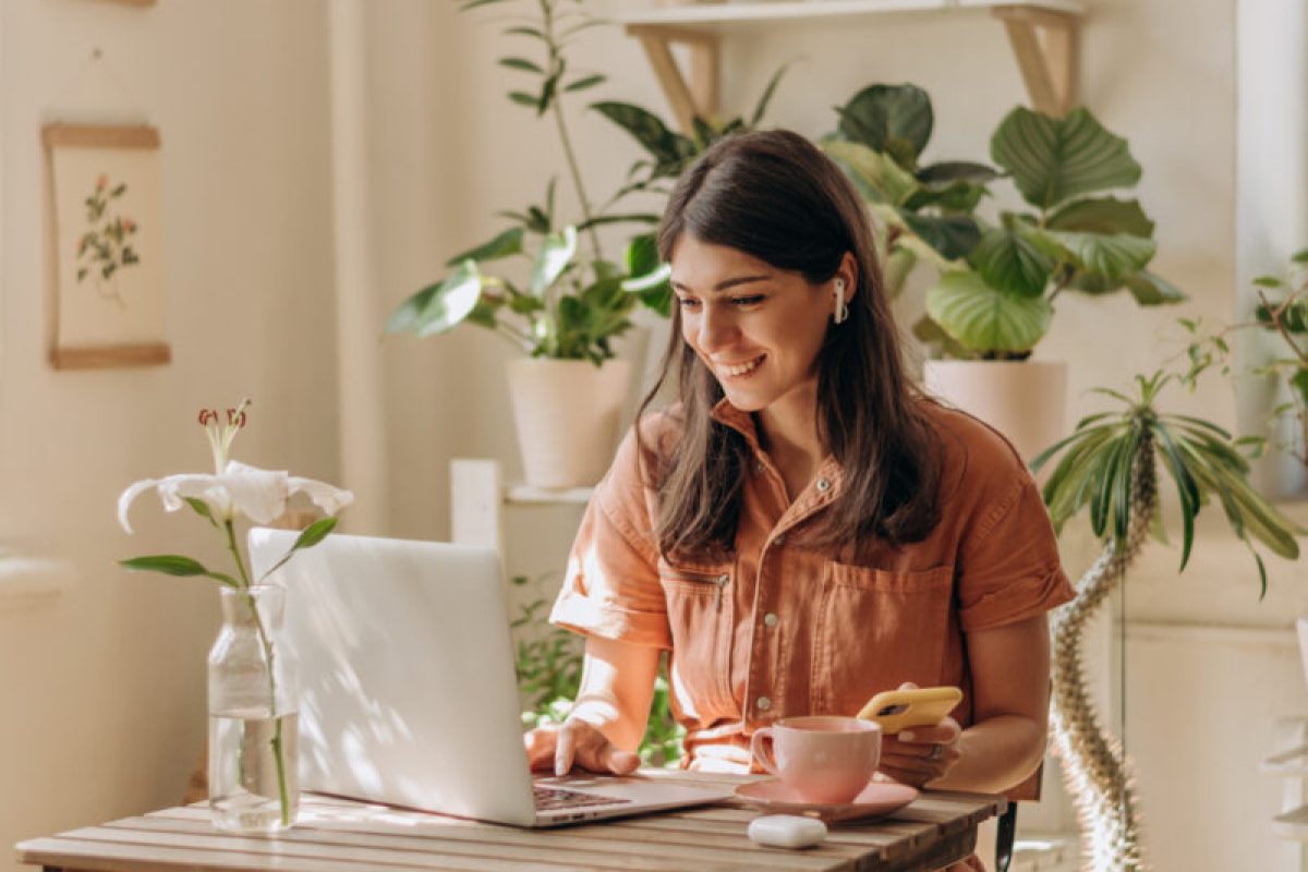 Positive young mixed race woman using a laptop and smartphone at home.Cozy home interior with indoor plants.Remote work, business,freelance,online shopping,e-learning,urban jungle concept Positive young mixed race woman using a laptop and smartphone at home.Cozy home interior with indoor plants.Remote work, business,freelance,online shopping,e-learning,urban jungle concept.
