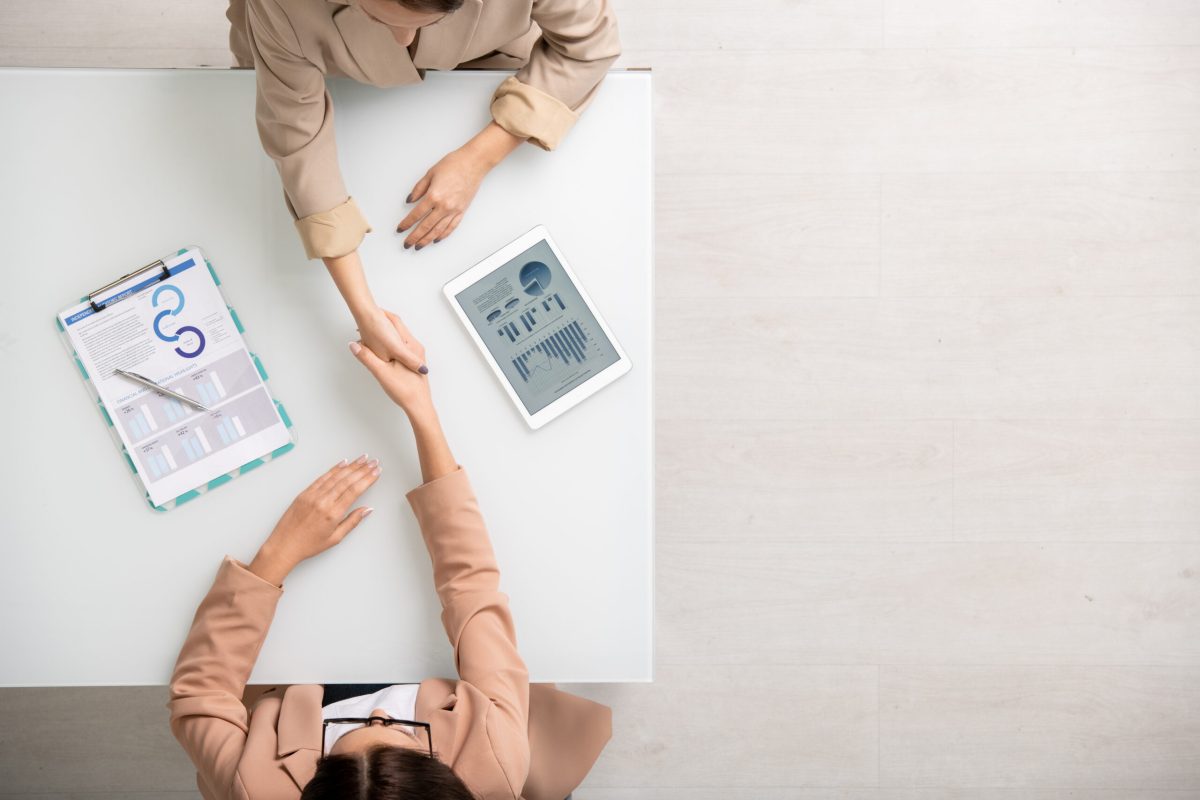 View of two young contemporary businesswomen shaking hands after negotiating View of two young contemporary businesswomen shaking hands over desk after making deal among financial information in tablet and on paper